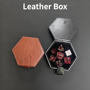K&ouml;nnte beinhalten: Zwei sechseckige Lederboxen, eine braune und geschlossene, die andere schwarze und offene, mit roten W&uuml;rfeln. Der Text "Leather Box" ist wei&szlig;. Die offene Box enth&auml;lt mehrere rote W&uuml;rfel.