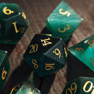 May include: A collection of polyhedral dice in shades of teal and dark green, with gold-colored numbers. The dice are arranged on a wooden surface, with some in focus and others blurred, creating a dynamic composition.
