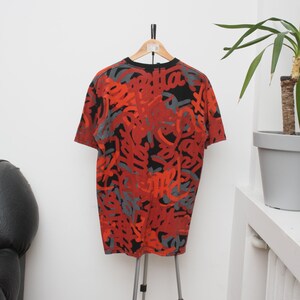 Puede incluir: Camiseta gr&aacute;fica roja, negra y gris colgada de un tr&iacute;pode. La camiseta de manga corta presenta un dise&ntilde;o abstracto repetitivo. La camiseta se muestra contra una pared blanca con una planta en una maceta gris.