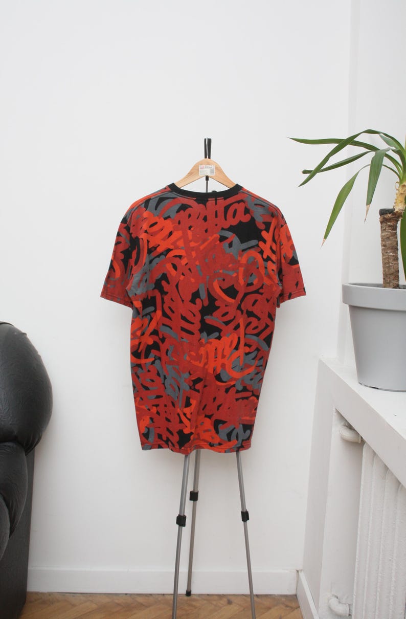 Puede incluir: Camiseta gr&aacute;fica roja, negra y gris colgada de un tr&iacute;pode. La camiseta de manga corta presenta un dise&ntilde;o abstracto repetitivo. La camiseta se muestra contra una pared blanca con una planta en una maceta gris.