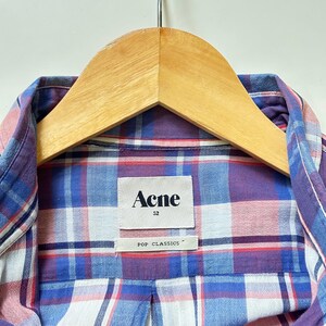 Peut inclure: Gros plan d'une chemise &agrave; carreaux dans les tons de bleu, violet et rose, suspendue &agrave; un cintre en bois. La chemise porte une &eacute;tiquette indiquant "Acne" et "POP CLASSICS". La chemise est de taille 52.