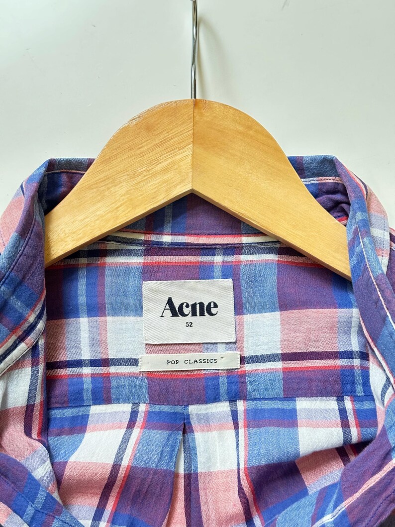 Peut inclure: Gros plan d'une chemise &agrave; carreaux dans les tons de bleu, violet et rose, suspendue &agrave; un cintre en bois. La chemise porte une &eacute;tiquette indiquant "Acne" et "POP CLASSICS". La chemise est de taille 52.