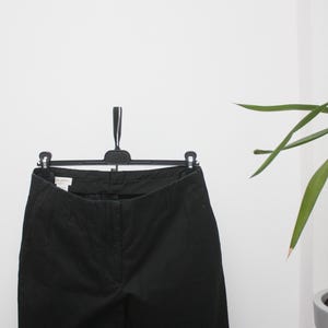 May include: A pair of black trousers hanging on a black clothes hanger. The trousers are made of a dark, solid fabric. A green plant is visible in the background, against a white wall. The trousers appear to be a classic style.