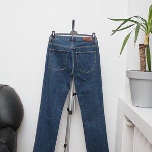 May include: Dark blue denim jeans hanging on a black hanger. The jeans have two back pockets and a brown leather patch. The jeans are displayed on a silver tripod stand against a white wall.