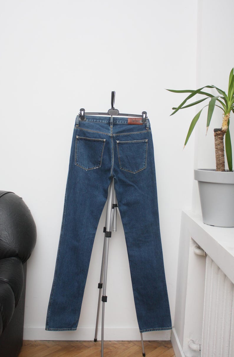 May include: Dark blue denim jeans hanging on a black hanger. The jeans have two back pockets and a brown leather patch. The jeans are displayed on a silver tripod stand against a white wall.
