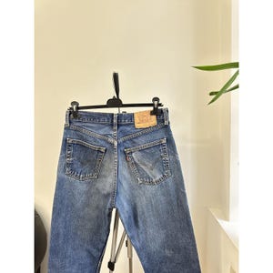 Könnte beinhalten: Blaue Jeans aus Denim mit klassischem Fünf-Taschen-Design. Die Jeans haben eine verwaschene Optik und einen Lederaufnäher am hinteren Bund. Die Gesäßtaschen haben ein charakteristisches Stichdesign. Die Jeans hängen an einem schwarzen Kleiderbügel.