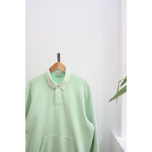 May include: A light green long-sleeved sweatshirt with a collared neckline and a front pocket. The sweatshirt is hanging on a wooden hanger against a white background. The collar and seams have a lighter green hue.