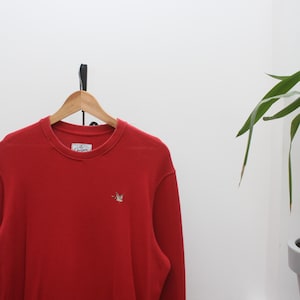 Puede incluir: Una sudadera roja de cuello redondo con un pequeño emblema de pájaro bordado en el pecho. La parte superior de manga larga está colgada de una percha de madera contra una pared blanca. La sudadera tiene un dobladillo acanalado.