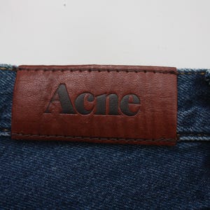 May include: Close-up of a pair of blue denim jeans with a brown leather patch. The patch is stitched onto the jeans and features the brand name "Acne" in black lettering.