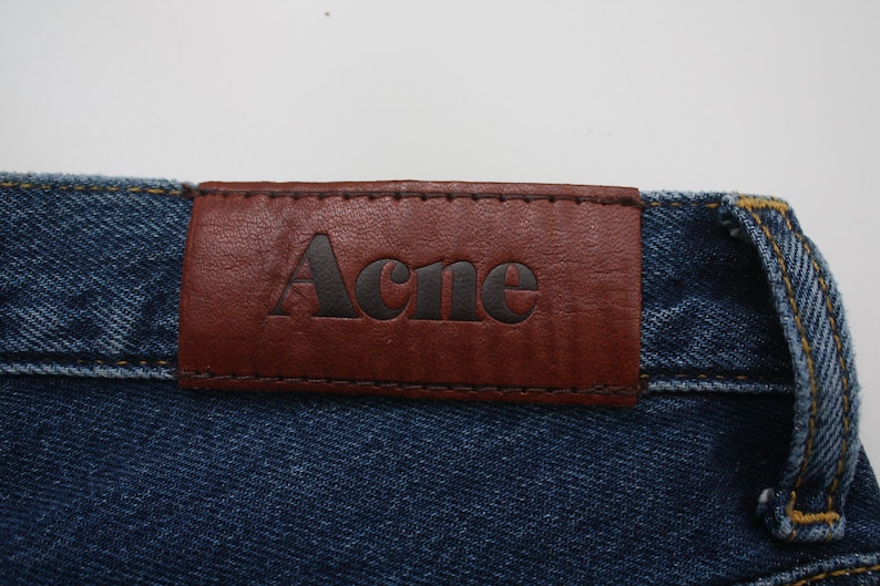 May include: Close-up of a pair of blue denim jeans with a brown leather patch. The patch is stitched onto the jeans and features the brand name "Acne" in black lettering.