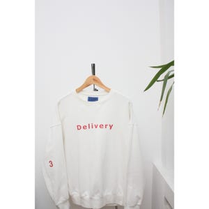 May include: A white sweatshirt with the word "Delivery" in red letters across the chest. The number "3" is printed in red on the left sleeve. The sweatshirt is hanging on a wooden hanger.