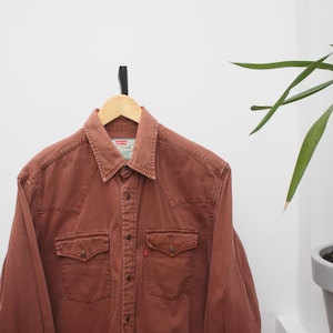 Op de afbeelding: Roodbruin overhemd met knoopsluiting, twee borstzakken en een kraag, hangend aan een houten hanger. Het overhemd heeft een klassieke westernstijl en een klein rood label op de zak.