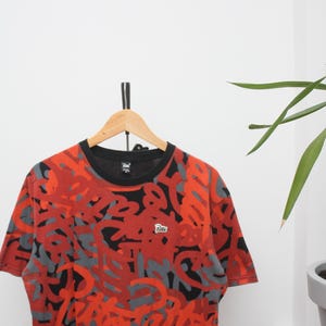 Puede incluir: Camiseta de manga corta con cuello negro y un vibrante estampado de formas abstractas rojas y grises. La camiseta est&aacute; colgada en una percha de madera sobre un fondo blanco. Un peque&ntilde;o logotipo es visible en la camiseta.