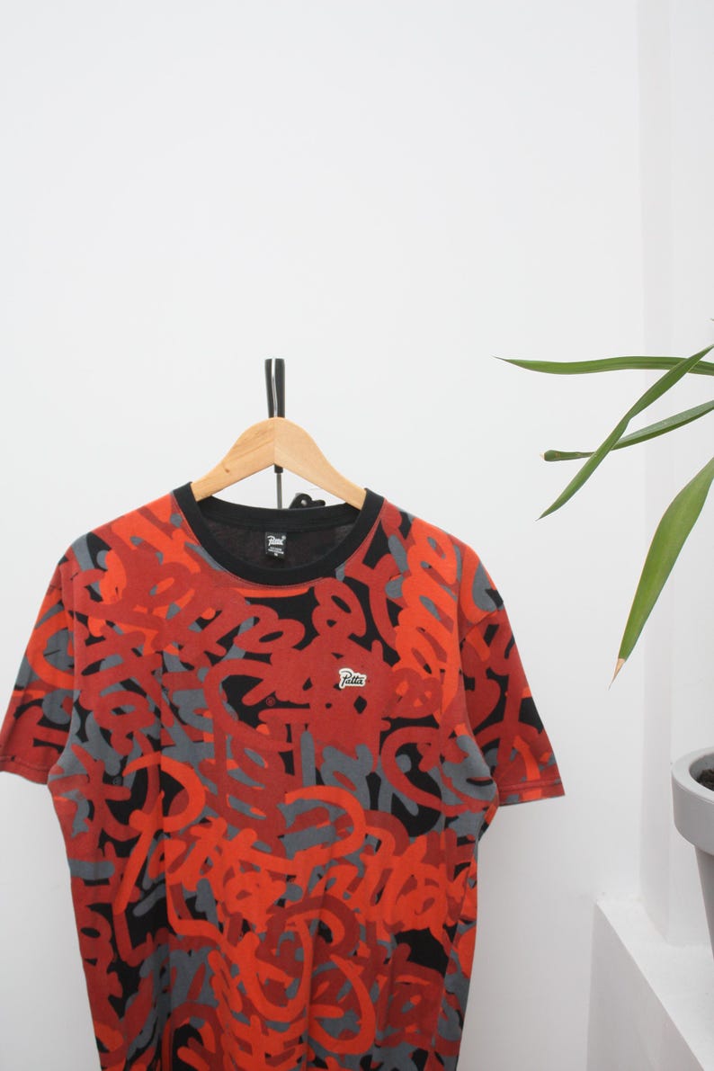 Puede incluir: Camiseta de manga corta con cuello negro y un vibrante estampado de formas abstractas rojas y grises. La camiseta est&aacute; colgada en una percha de madera sobre un fondo blanco. Un peque&ntilde;o logotipo es visible en la camiseta.