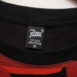 Puede incluir: Primer plano de una sudadera negra de cuello redondo con un estampado rojo y gris. La etiqueta negra dice "Patta EST.2004 AMSTERDAM M". La sudadera tiene un cuello acanalado.