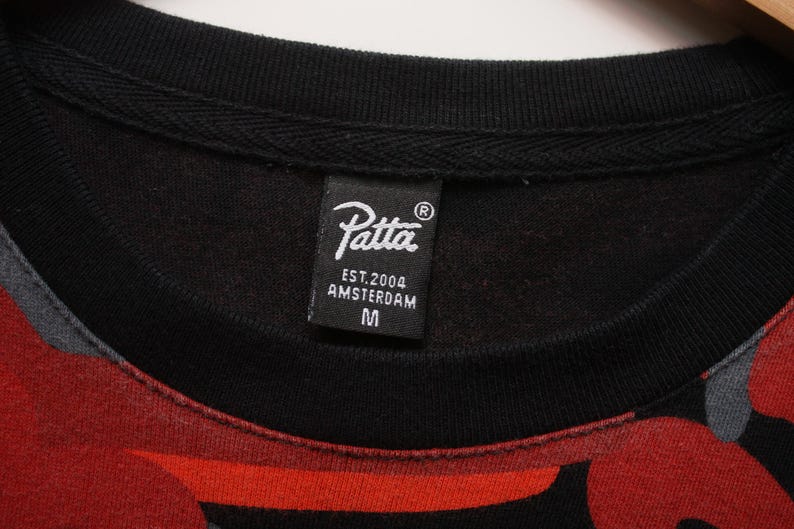 Puede incluir: Primer plano de una sudadera negra de cuello redondo con un estampado rojo y gris. La etiqueta negra dice "Patta EST.2004 AMSTERDAM M". La sudadera tiene un cuello acanalado.