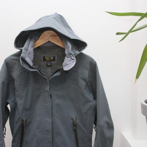 Puede incluir: Chaqueta gris con capucha, cierre de cremallera completo y dos bolsillos delanteros. La chaqueta está colgada de una percha de madera. La chaqueta tiene la marca "Mountain Hardwear" en el pecho.