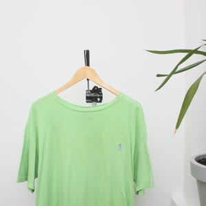 May include: A pale green short-sleeved t-shirt with a small blue logo on the chest. The shirt hangs on a wooden hanger against a white wall. A plant is visible in the background.