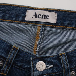 May include: Close-up of blue denim jeans with the brand name "Acne" on a white tag. The jeans have a button closure and detailed stitching in a contrasting yellow-brown thread.