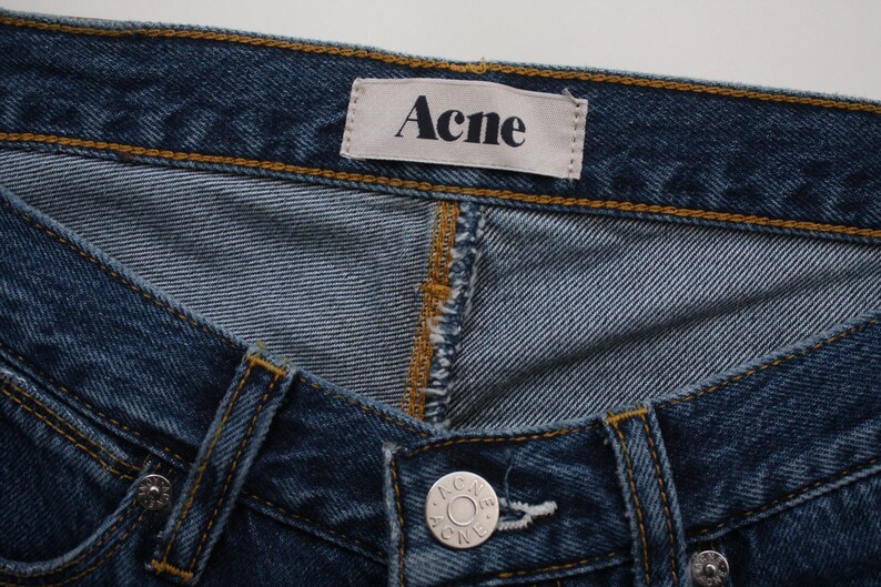 May include: Close-up of blue denim jeans with the brand name "Acne" on a white tag. The jeans have a button closure and detailed stitching in a contrasting yellow-brown thread.