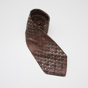 May include: Rolled brown tie with a repeating floral pattern in shades of brown and tan. The tie is made of a shiny fabric and is displayed on a white background. The tie is a classic accessory.