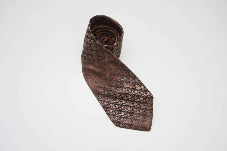 May include: Rolled brown tie with a repeating floral pattern in shades of brown and tan. The tie is made of a shiny fabric and is displayed on a white background. The tie is a classic accessory.