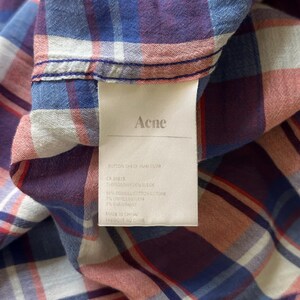 Peut inclure: Gros plan sur une chemise &agrave; carreaux dans des tons de bleu, rouge et violet. La chemise porte une &eacute;tiquette avec le nom "Acne" et d'autres informations. Le tissu semble &ecirc;tre du coton. La chemise est pli&eacute;e, montrant la texture et le motif.