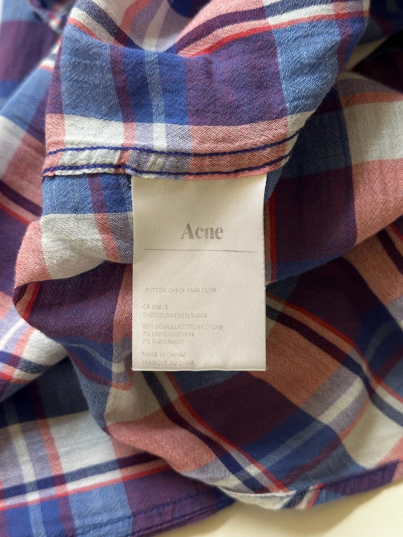Peut inclure: Gros plan sur une chemise &agrave; carreaux dans des tons de bleu, rouge et violet. La chemise porte une &eacute;tiquette avec le nom "Acne" et d'autres informations. Le tissu semble &ecirc;tre du coton. La chemise est pli&eacute;e, montrant la texture et le motif.
