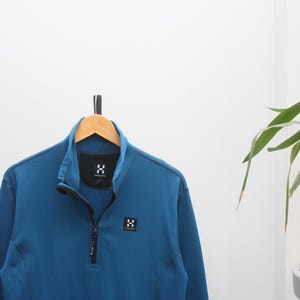 Könnte beinhalten: Türkisblaue Fleecejacke mit Reißverschluss und hohem Kragen, hängt an einem Holzbügel. Die Jacke hat einen schwarzen Reißverschluss und kleine weiße Logoaufnäher. Die Jacke ist aus einem weichen, warmen Material gefertigt.