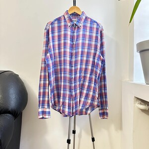 Peut inclure: Une chemise boutonn&eacute;e &agrave; manches longues avec un motif &agrave; carreaux bleus, roses et blancs. La chemise est pr&eacute;sent&eacute;e sur un cintre en bois et un tr&eacute;pied. La chemise a un col boutonn&eacute; et des poignets.