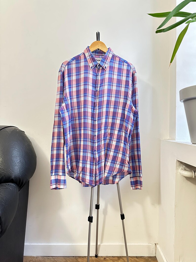 Peut inclure: Une chemise boutonn&eacute;e &agrave; manches longues avec un motif &agrave; carreaux bleus, roses et blancs. La chemise est pr&eacute;sent&eacute;e sur un cintre en bois et un tr&eacute;pied. La chemise a un col boutonn&eacute; et des poignets.