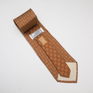 Op de afbeelding: Bruine zijden stropdas met een herhalend patroon van goudkleurige schakels en stippen. Het label luidt "GIANNI VERSACE". Deze stropdas is gemaakt van een luxueus materiaal en is geschikt voor formele gelegenheden. Een klassiek accessoire.