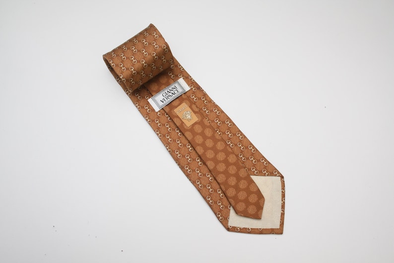 Op de afbeelding: Bruine zijden stropdas met een herhalend patroon van goudkleurige schakels en stippen. Het label luidt "GIANNI VERSACE". Deze stropdas is gemaakt van een luxueus materiaal en is geschikt voor formele gelegenheden. Een klassiek accessoire.