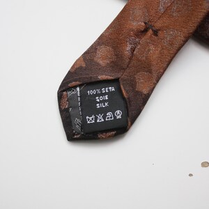 May include: Close-up of a brown silk tie with a subtle pattern. The tie's label reads "100% Seta Soie Silk" and includes care symbols. The tie is a classic accessory, suitable for formal or business attire.