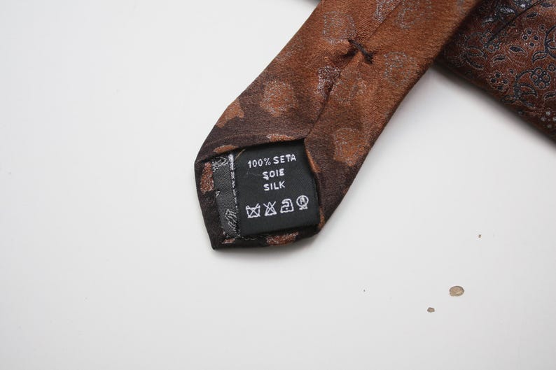 May include: Close-up of a brown silk tie with a subtle pattern. The tie's label reads "100% Seta Soie Silk" and includes care symbols. The tie is a classic accessory, suitable for formal or business attire.