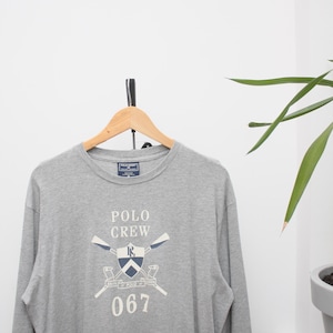 May include: A grey long-sleeved shirt displaying the text "POLO CREW" and a crest design with crossed oars and the number "067". The shirt is suspended on a wooden hanger.