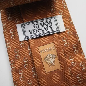 Op de afbeelding: Een bruine zijden stropdas met een herhalend patroon van witte paardenbitjes en diagonale strepen. De stropdas heeft het label "GIANNI VERSACE" en een kleiner label met het Versace-logo.
