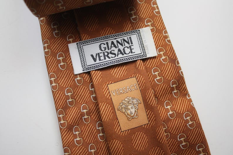 Op de afbeelding: Een bruine zijden stropdas met een herhalend patroon van witte paardenbitjes en diagonale strepen. De stropdas heeft het label "GIANNI VERSACE" en een kleiner label met het Versace-logo.