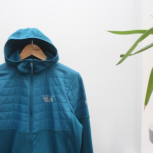 Pode incluir: Casaco com capuz azul-petróleo com uma secção superior acolchoada e um fecho de correr completo. O casaco apresenta o logótipo "Mountain Hardwear" no peito. O capuz está preso e o casaco está pendurado num cabide de madeira.