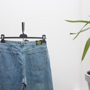 Puede incluir: Vaqueros de mezclilla azul claro colgando de una percha negra. Los vaqueros tienen dos bolsillos traseros y un pequeño parche verde y amarillo en la parte trasera. Los vaqueros se exhiben contra una pared blanca.