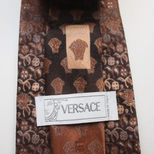 May include: A brown Versace tie with a repeating pattern of floral and Medusa head motifs. The tie features a central panel with the Versace logo and a label that reads "Made in Italy".