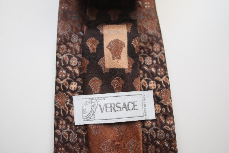 May include: A brown Versace tie with a repeating pattern of floral and Medusa head motifs. The tie features a central panel with the Versace logo and a label that reads "Made in Italy".