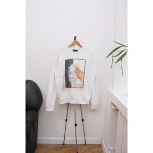 May include: White sweatshirt with a graphic print of a person's face, half in black and white, with a hand covering the eye. The sweatshirt is hanging on a wooden hanger on a tripod stand.