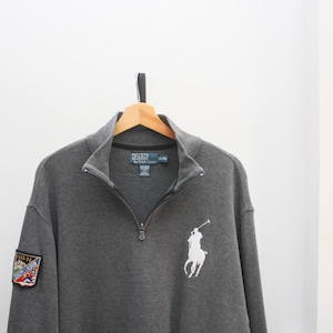 May include: A charcoal gray Polo by Ralph Lauren quarter-zip sweatshirt. The sweatshirt features a white embroidered polo player logo on the chest and a patch on the left sleeve. The collar is black, and the zipper is silver.