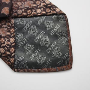May include: Close-up of a brown silk tie with a floral pattern in shades of brown and tan. The interior lining is black with the Versace logo in white. The tie is folded over, revealing the lining.
