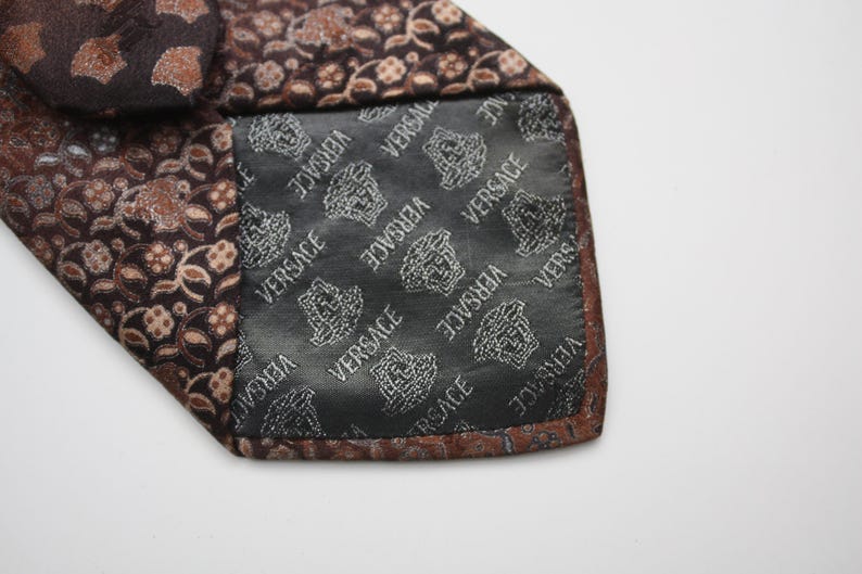 May include: Close-up of a brown silk tie with a floral pattern in shades of brown and tan. The interior lining is black with the Versace logo in white. The tie is folded over, revealing the lining.