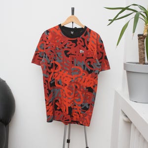 Puede incluir: Una camiseta estampada en rojo, negro y gris cuelga de una percha de madera. La camiseta tiene cuello redondo y mangas cortas. Un peque&ntilde;o logotipo es visible en el pecho. La camiseta se exhibe en un tr&iacute;pode plateado.