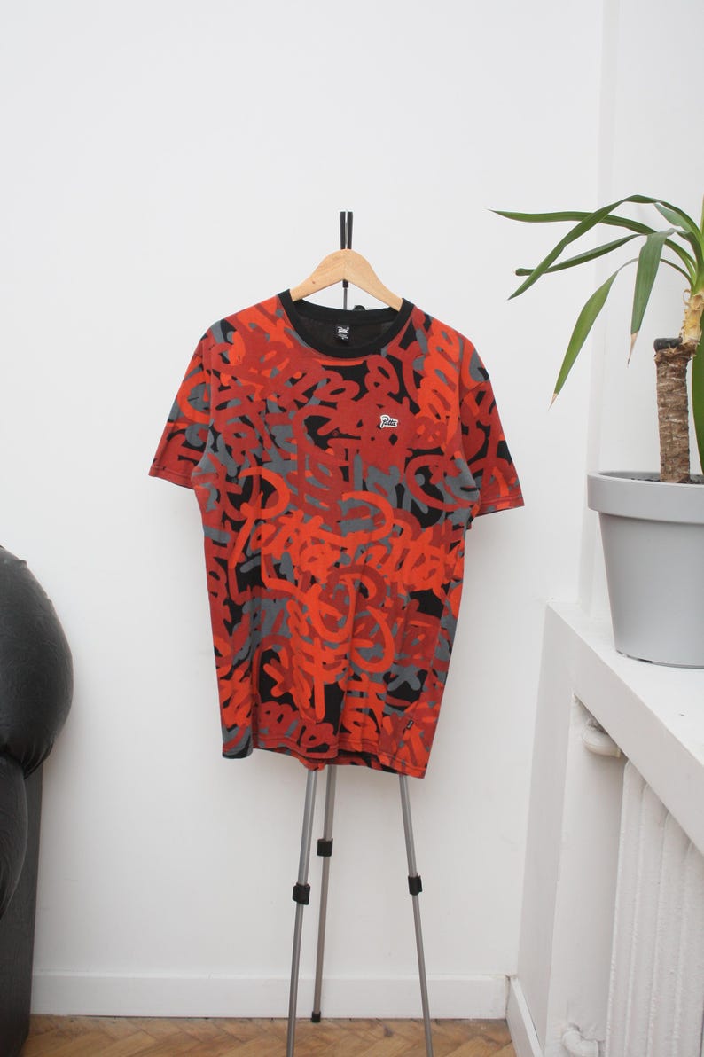 Puede incluir: Una camiseta estampada en rojo, negro y gris cuelga de una percha de madera. La camiseta tiene cuello redondo y mangas cortas. Un peque&ntilde;o logotipo es visible en el pecho. La camiseta se exhibe en un tr&iacute;pode plateado.