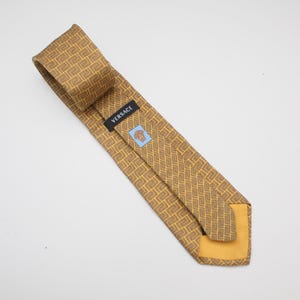 Puede incluir: Corbata Versace dorada con un patrón geométrico repetitivo. La etiqueta negra lleva el nombre de la marca "Versace" y un cuadrado azul con un logotipo. La corbata está hecha de un material similar a la seda.