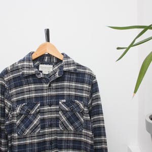 May include: A blue and white checked flannel shirt with two chest pockets, hanging on a wooden hanger. The shirt has a button-down front and a collared neckline. The fabric appears to be soft and comfortable.